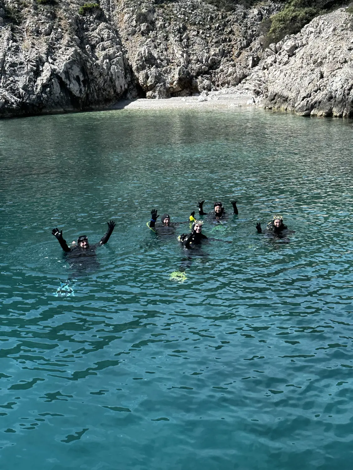 Eine Gruppe von Menschen beim Tauchen oder Schnorcheln in einem klaren Gewässer, wahrscheinlich einer Bucht, mit einer felsigen Küste im Hintergrund. Von Hang-Loose-Diving d.o.o.